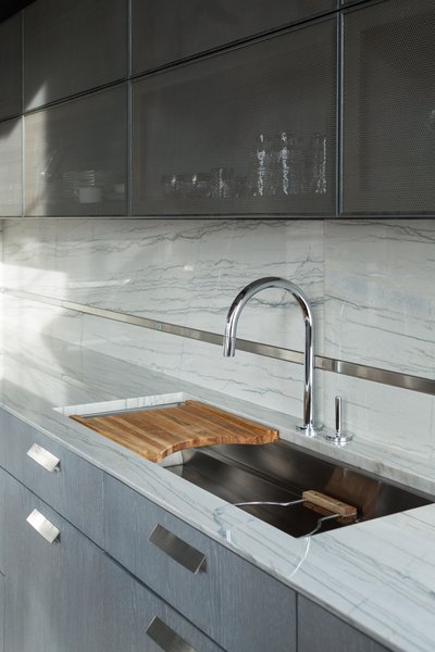 This kitchen pairs custom-stained oak cabinetry and white Macaubas quartzite countertops with perforated metal upper cabinets. These distinct components come together at the backsplash, which blends quartzite with a strip of stainless steel that houses electrical outlets and matches the minimalist, sleek faucet.
